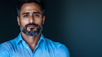 Middle-aged South Asian man with salt and pepper beard wearing blue collared shirt against dark background, confident expression and professional headshot style portrait.