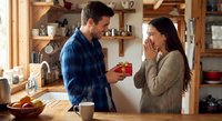 Young Caucasian man giving red gift box to surprised brunette woman in cozy kitchen during romantic moment together at home.