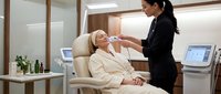 Professional Asian female aesthetician performing facial treatment on relaxed Caucasian woman client in modern luxury spa clinic with advanced equipment.