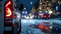 Car taillight glowing red on snowy city street at night with blurred traffic lights and urban bokeh background creating atmospheric winter driving scene.