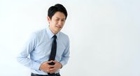 Young Asian businessman in blue shirt and tie holding stomach with painful expression due to digestive issues or food poisoning on white background.