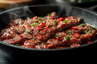Juicy beef strips stir-fried in dark sauce with red chili peppers and green onions on black pan, steam rising, close up view for restaurant menu or cookbook.