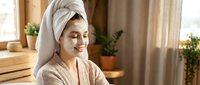 Young Asian woman with white towel on head applying facial mask in bright bathroom for skincare routine and spa treatment at home.