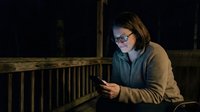 Middle-aged Caucasian woman wearing glasses and casual cardigan using smartphone on wooden porch at night for communication, work, or social media browsing.