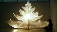 Illuminated translucent leaf sculpture with intricate vein patterns creates dramatic lighting effect as silhouetted figure observes artistic installation.