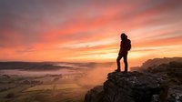 Silhouette of hiker standing on rocky cliff overlooking misty valley at sunset with dramatic orange and pink sky, adventure and exploration concept.