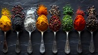 Colorful aromatic spices in vintage silver spoons on dark slate background, overhead view of turmeric, black pepper, salt, paprika, and dried herbs arranged in row.