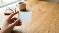 Hand holding blank white card on wooden desk with coffee cup and eyeglasses, suitable for business branding and stationery mockups.