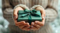 Female hands holding small green gift box with emerald ribbon bow, presenting special occasion present in cozy sweater setting.