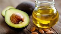 Fresh avocado halves with pit and golden oil in glass jar surrounded by almonds on wooden surface for healthy cooking and natural skincare routines.
