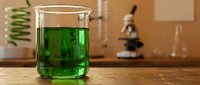 Green liquid in glass beaker on wooden laboratory table with microscope and scientific equipment in background for chemistry research and education.