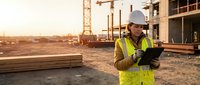 Construction worker in safety gear using tablet at building site during golden hour sunset for project management and digital planning.