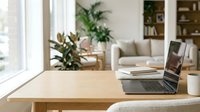 Laptop on a wooden desk in a bright modern home office with cozy living room in background, natural light, remote work concept.