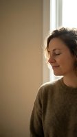Peaceful young woman with closed eyes enjoying warm sunlight by window wearing cozy sweater in minimalist home interior for wellness and mindfulness concepts.