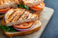 Grilled chicken breast sandwich with fresh tomatoes, lettuce and red onion on artisan bread served on wooden cutting board for healthy meal preparation.