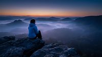 Hiker in blue jacket sits on rocky mountain peak at dawn, gazing over misty forested valleys and distant mountains during golden sunrise hour.