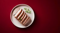 Sliced roasted pork tenderloin with herbs on white plate against burgundy background for culinary presentations and food photography.