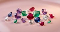 Colorful collection of precious gemstones including diamonds, emeralds, rubies, sapphires and amethysts scattered on soft pink surface for jewelry design.