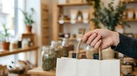 Person holding white paper shopping bag in modern retail store with wooden shelves and potted plants, suitable for e-commerce and retail marketing.