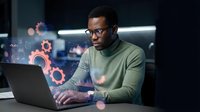 Young African American male software developer working on laptop with floating digital gears and technology elements in modern office environment.