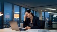 Stressed businesswoman working late at night in modern office with city lights, experiencing work burnout and fatigue at laptop computer.