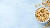 Crunchy honey-flavored cereal rings scattered around white ceramic bowl on light blue background. Top view of breakfast cereals with copy space.