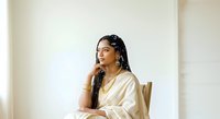 Young South Asian woman with long braided hair and traditional jewelry, wearing a cream-colored sari, seated thoughtfully against a neutral background for portrait and lifestyle use.