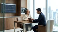 Middle-aged Caucasian woman in business attire and South Asian man in dark suit having professional discussion at wooden desk in modern high-rise office with city skyline view, stock market display visible.