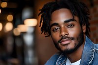 Young African American man with dreadlocks and denim jacket smiling confidently against blurred warm bokeh background, expressing joy and authenticity in casual setting.
