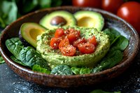 Fresh avocado guacamole dip garnished with cherry tomatoes and spinach leaves in rustic ceramic bowl, served with sliced avocado on dark background.