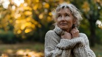 Senior woman with curly blonde hair wearing cozy knit sweater in autumn park, looking thoughtfully upward with serene expression against golden foliage background.