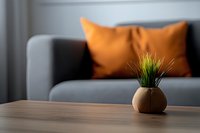 Small decorative grass plant in round ceramic pot on wooden table with gray sofa and orange cushion in blurred background creating minimalist home atmosphere.
