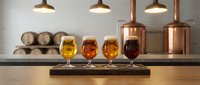 Four craft beer glasses with different colored ales arranged on wooden board in modern brewery with copper equipment and pendant lighting for tasting.
