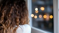 Young woman with curly hair looking through window at night with bokeh lights in background, creating atmospheric mood for lifestyle and emotional concepts.