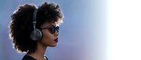 Young African American woman with curly afro hair wearing wireless headphones and sunglasses against blue sky background for music and lifestyle content.