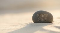 Young adult male, Caucasian ethnicity, depicted as a businessman in casual attire, standing on sandy beach holding price hike sign carved into stone rock, symbolizing economic inflation and rising costs for business and finance concepts.
