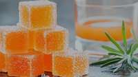 Orange jelly cubes covered in sugar crystals stacked near glass with juice and succulent plant, fresh citrus dessert sweets on gray background.