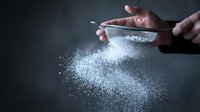 Hands sifting white flour through fine mesh strainer creating powder cloud on dark surface for baking and cooking preparation.