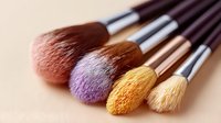 Professional makeup brushes in pink, purple, yellow and white colors with powder residue on bristles against neutral beige background, macro shot.