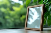 Empty wooden photo frame standing on windowsill with green foliage background and soft natural lighting creating shadow patterns on glass surface.