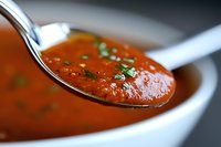 Fresh homemade tomato soup garnished with chopped herbs in white ceramic bowl, close-up view of spoon lifting creamy red soup showing texture and depth.