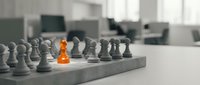 Young professional woman of mixed ethnicity in business attire leading a team meeting in modern office setting, symbolizing leadership and strategic planning through chess metaphor with orange pawn standing out among gray pieces.