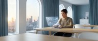 Young professional man working on laptop in modern office with city skyline view through large windows during golden hour lighting.