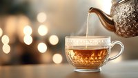 Hot tea pouring from vintage silver teapot into transparent glass cup with ornate pattern against blurred warm bokeh lights background, creating cozy atmosphere.
