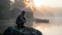 Man in hoodie crouching on rock by misty lake at sunrise with wooden boat and cabin in background creating peaceful morning atmosphere.