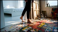Artist standing barefoot on colorful paint-splattered floor in bright studio with mountain view, creative workspace for art professionals and designers seeking inspiration and workspace imagery.