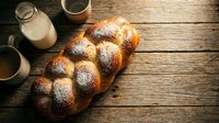 Fresh braided challah bread with powdered sugar on rustic wooden table with milk and coffee cups for breakfast bakery display.