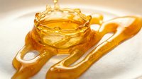Golden honey dripping from wooden dipper and spreader on white surface, sweet liquid food ingredient for cooking and baking applications.