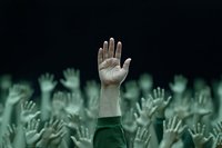 Raised hands in crowd, one prominent hand in focus against blurred background of multiple raised arms in green tonal atmosphere. Concept of democracy, voting, unity.