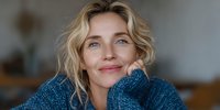 Smiling middle-aged Caucasian woman with blonde wavy hair wearing blue knit sweater, resting chin on hand in cozy home setting.